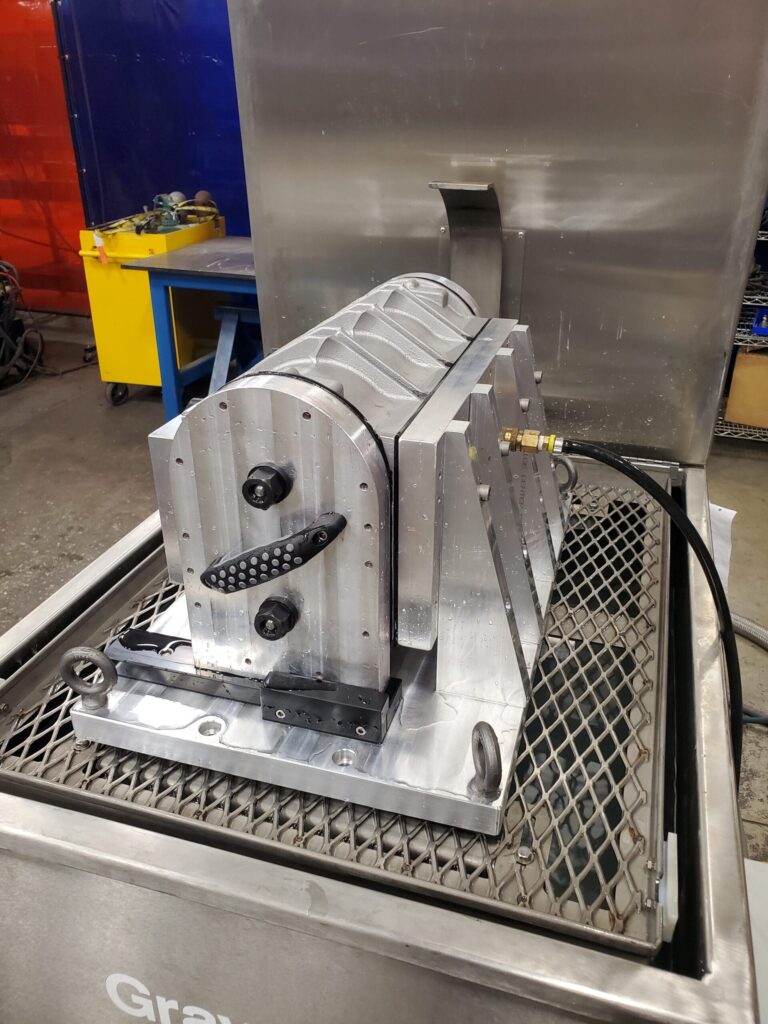 A metal fixture or jig placed on a grated surface, featuring machined components, a handle, and a black hose connected to it.