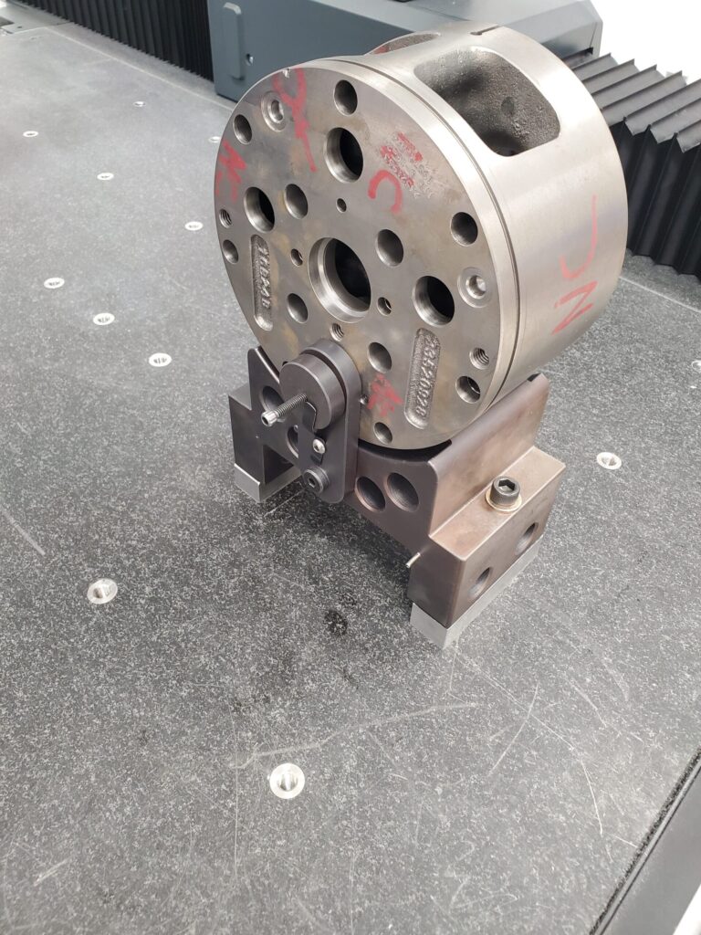 The image shows a metal fixture or jig mounted on a granite surface plate. The fixture appears to be used for holding or aligning parts during machining or inspection processes. It has multiple holes and markings, including some red markings, and is secured with bolts to the base.