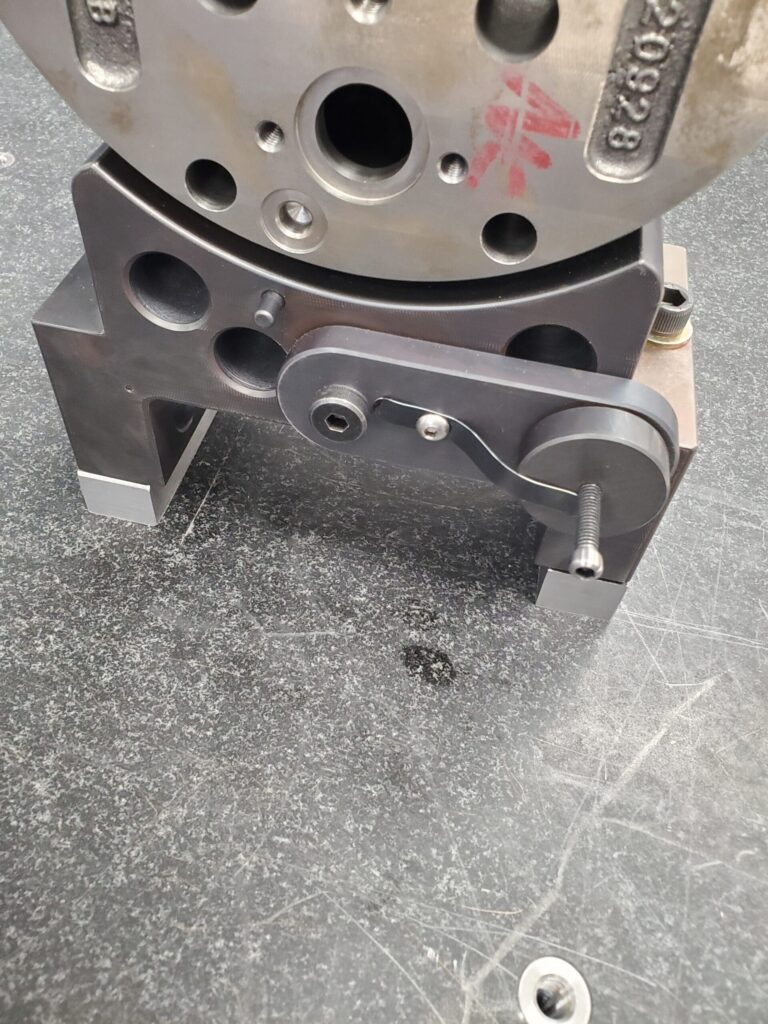 The image shows a metal mechanical component mounted on a black surface. The component appears to be part of a larger machine or assembly, featuring several circular holes and a lever mechanism with a handle.