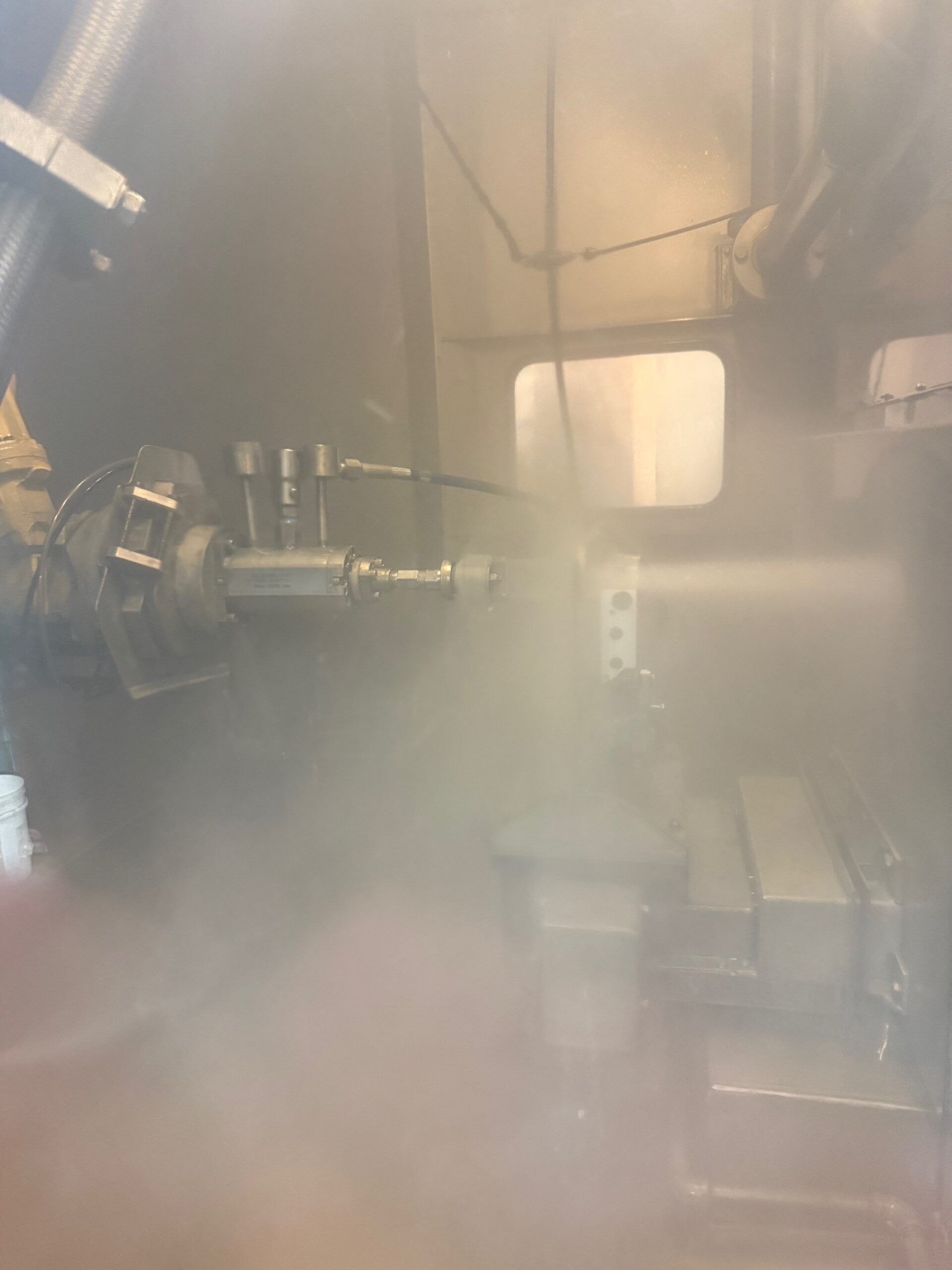 A misty or foggy environment inside an industrial or mechanical setting, featuring a machine with various components including pipes, valves, and possibly a rotating mechanism. The visibility is low due to the mist or fog present in the scene.