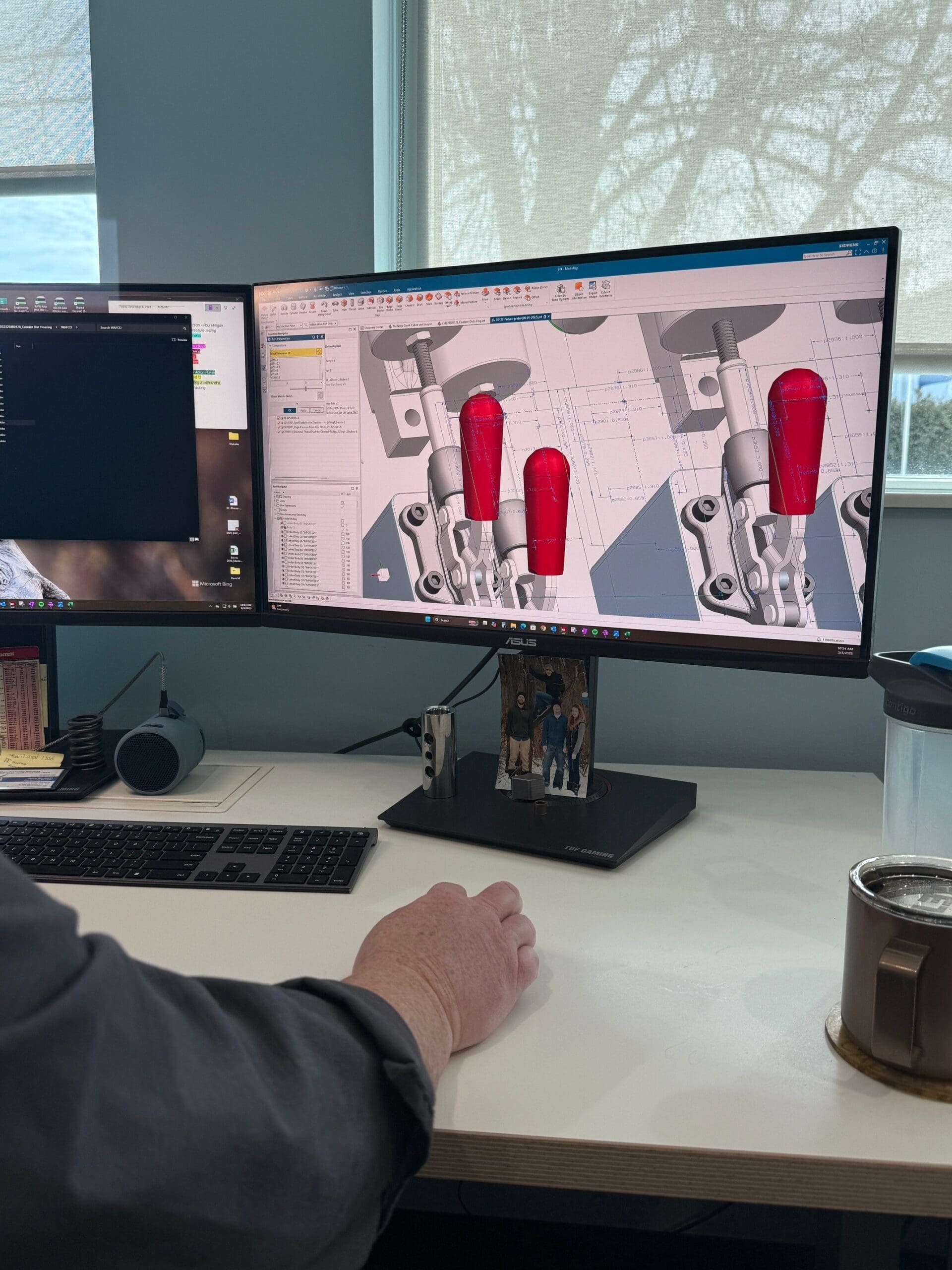 An engineer's workspace featuring a large computer monitor displaying a 3D CAD model of mechanical components. The model includes several red and gray parts, likely representing different materials or components in an assembly.