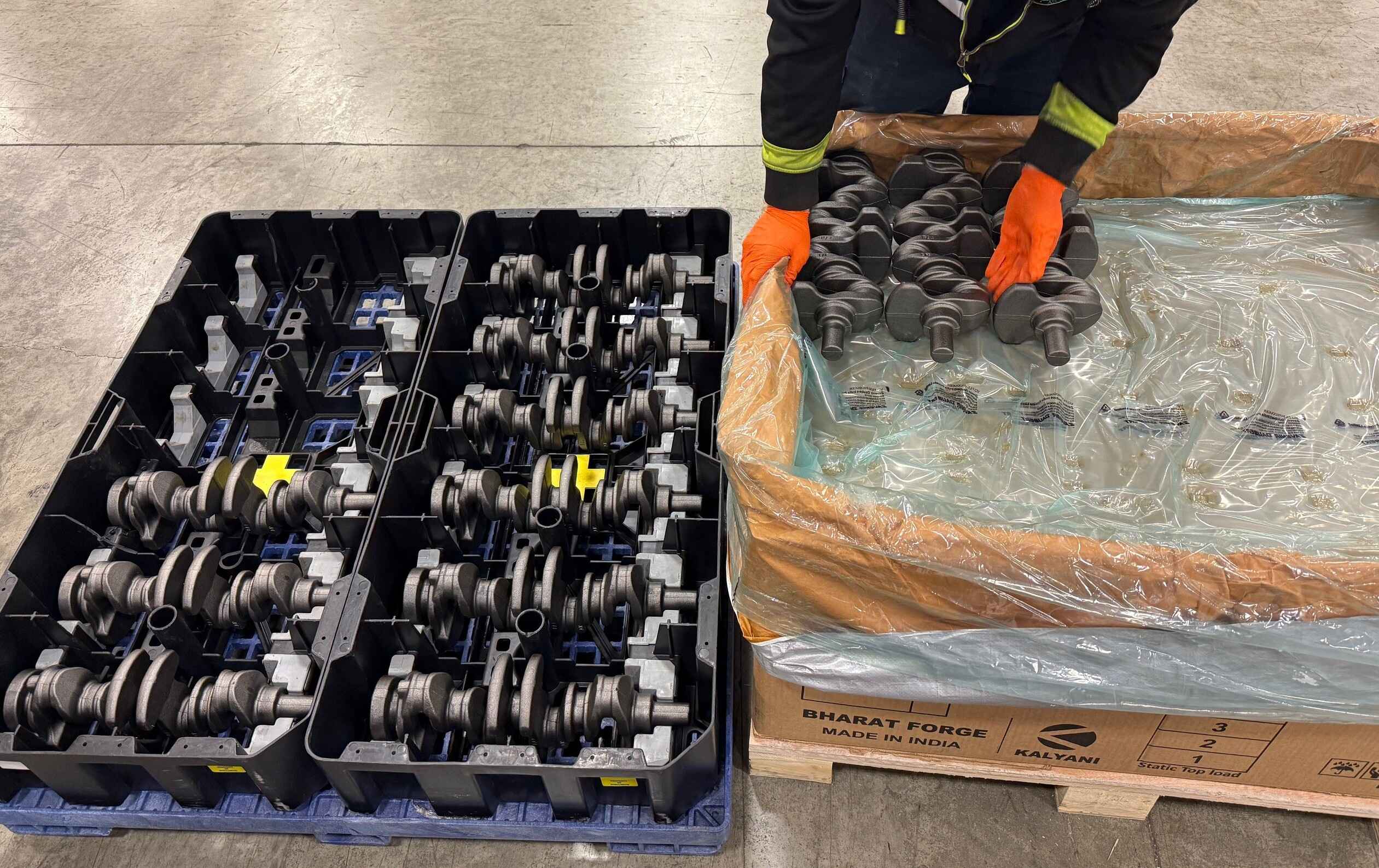 A person handling metal crankshafts, placed in a plastic container and a wooden crate labeled 'BHARAT FORGE' and 'MADE IN INDIA.' The person is wearing orange gloves and appears to be organizing or inspecting the crankshafts.