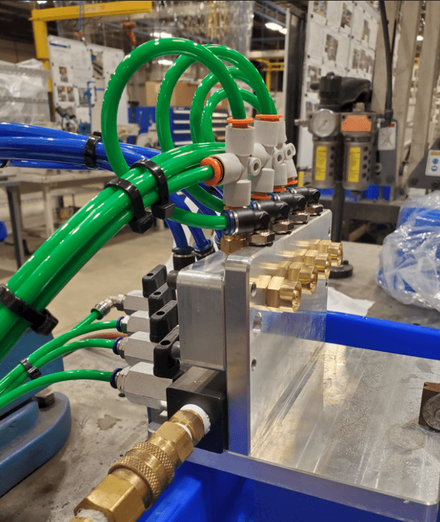 A mechanical setup in an industrial or laboratory environment featuring green and blue hoses connected to a metallic block with brass fittings and valves.