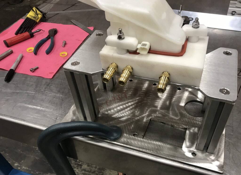 The image shows a metal workbench with various tools and components. There is a white plastic component mounted on a metal fixture with three brass fittings protruding from it. The fixture is secured to the workbench with a C-clamp. On the left side of the image, there are tools including pliers, markers, and screws placed on a red cloth.