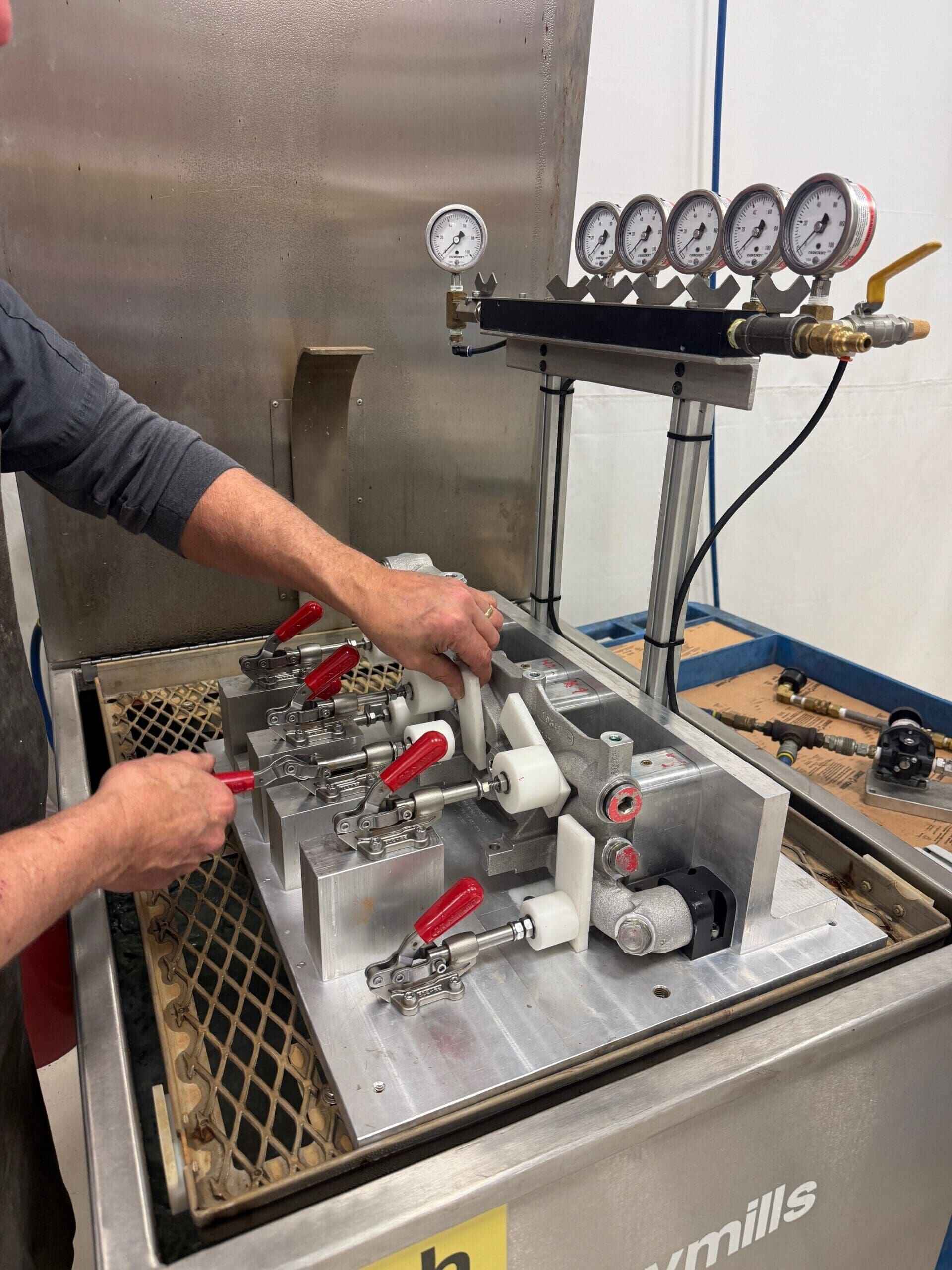 A technician working with industrial equipment labeled 'Wymills,' featuring several gauges, valves, and red-handled levers. The technician is adjusting or assembling parts on the machine, which has four pressure gauges mounted on a horizontal bar at the top.