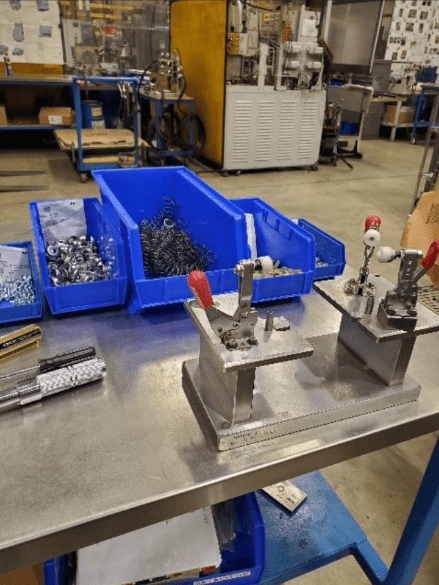 The image shows a workspace in a mechanical or manufacturing workshop. There are several blue bins on the table containing various metal parts, such as screws and springs. In the foreground, there are two metal fixtures with red handles mounted on a metallic surface. The background includes more workbenches, tools, and machinery.