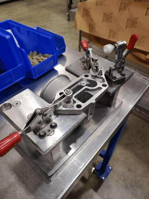 The image shows a metal fixture or jig on a workbench, likely used for holding and securing parts during machining or assembly. The fixture has two red-handled clamps and additional components to hold the part in place. There are blue bins in the background containing small parts, and a piece of brown paper with text is partially visible on the right side of the image.