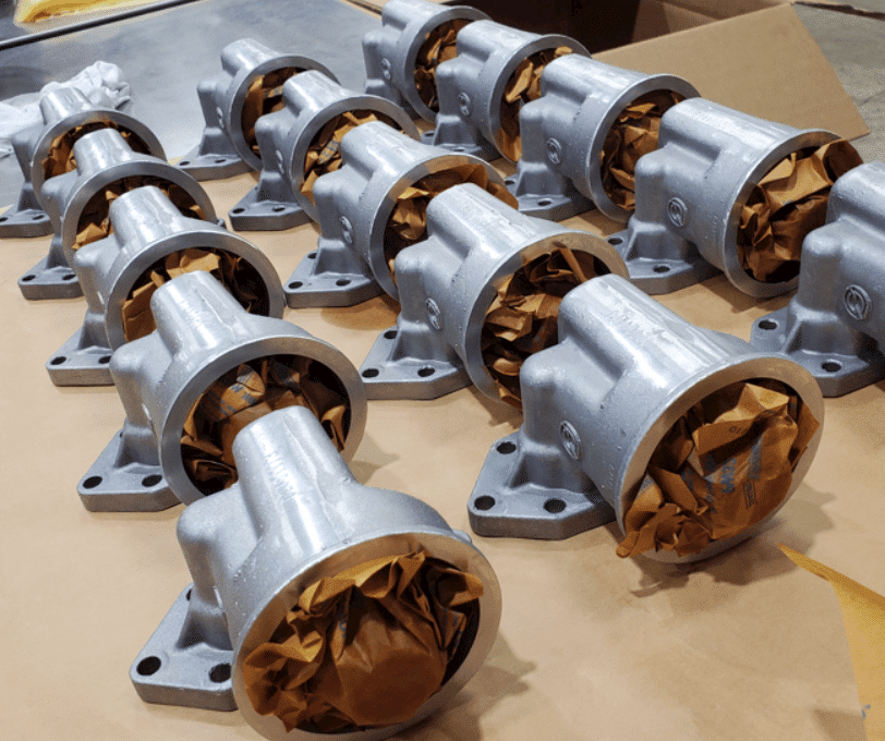 The image shows a series of metallic components arranged in rows on a surface. Each component appears to be cylindrical with flanges and mounting holes, and the openings are filled with crumpled brown paper.