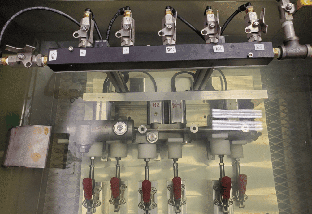 The image shows a mechanical setup with multiple valves and levers. There are five labeled valves at the top (W1, W2, K4, K1, K5) connected to a black bar. Below this bar are two additional components labeled H2 and K-1. At the bottom of the image, there are five red-handled levers attached to cylindrical components. The background has a patterned surface.