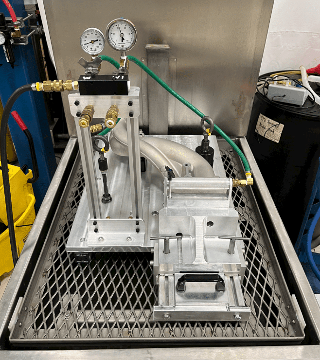 The image shows a precision fixture and tooling setup used for engine component testing. The setup includes a metal frame with two pressure gauges mounted on top, connected to various valves and hoses. Adjacent to the frame is a custom fixture designed to hold an engine manifold securely in place during testing. The entire assembly is situated on a metal mesh platform within an industrial test stand, surrounded by additional equipment and tools.