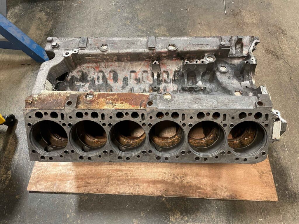 A used and worn engine block with six large cylindrical openings for pistons, placed on a wooden board. The surface is rusty and corroded, with various bolt holes and mounting points visible.
